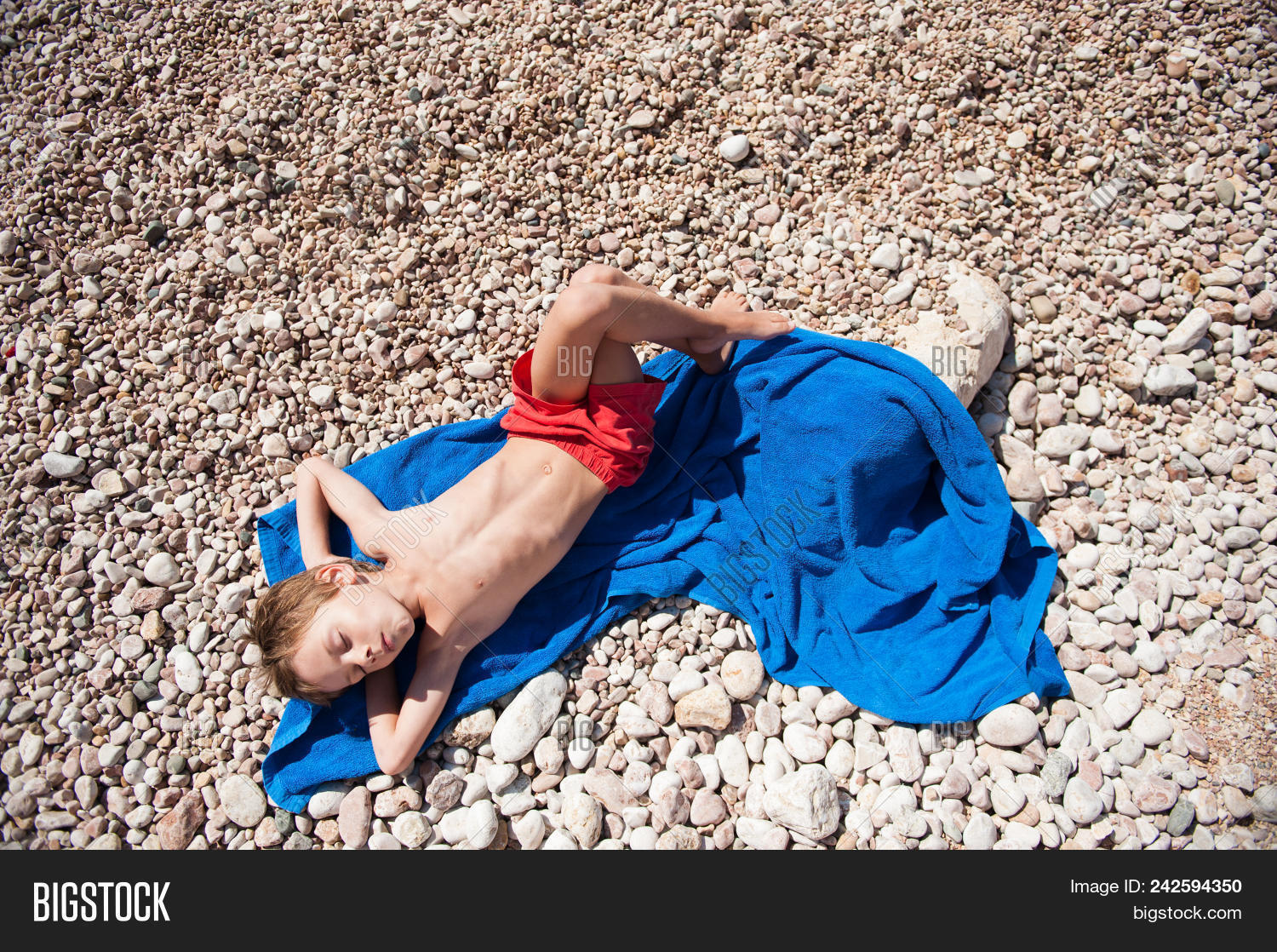 Healthy Little Boy In Red Shorts Lying On Blue Towel On Beach In Hot