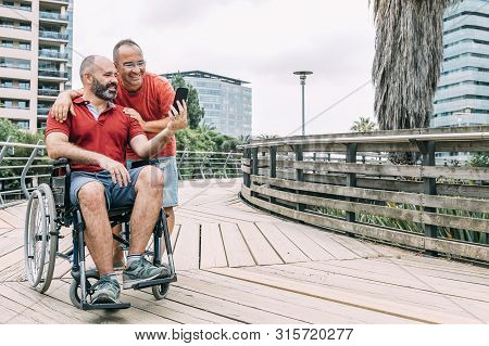 Disabled Man In Wheelchair Making A Selfie With His Phone With A Friend During A Walk, Concept Of Fr