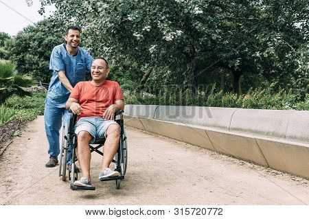 Disabled Man In Wheelchair Laughing During A Walk With His Nurse At Park, Concept Of Medical Care An