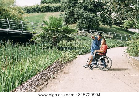 Caretaker Talking With His Disabled Patient In A Wheelchair During A Walk, Concept Of Medical Care A