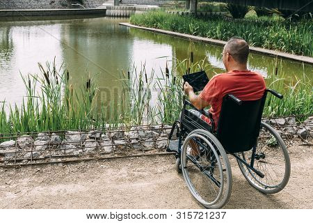 Disabled Man In Wheelchair Having Fun While Resting Using A Tablet Computer At Park, Concept Of Tech