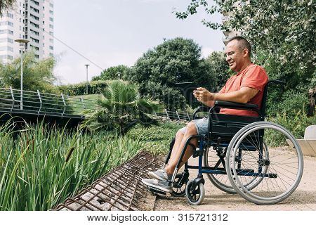 Handicapped Man In Wheelchair Having Fun While Working Using A Tablet Computer At Park, Concept Of T