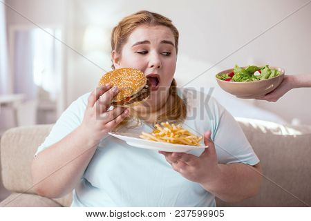 Junk Food. Fat Young Woman Eating Unhealthy Food While Thinking About Nice Green Salad